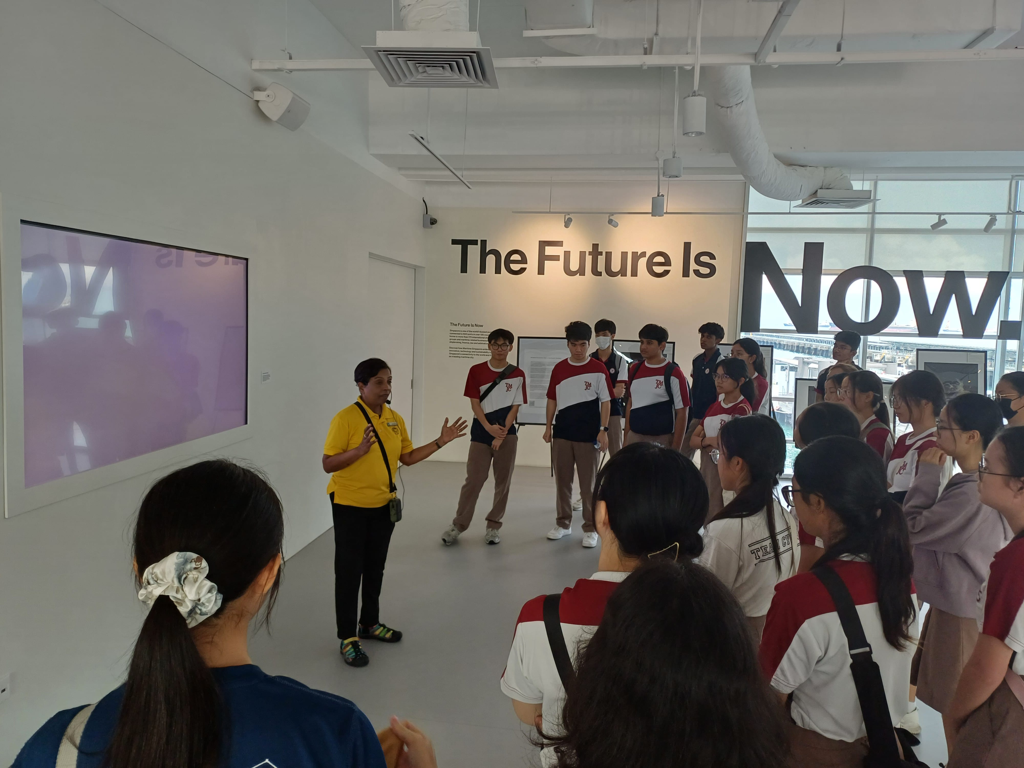 A group of students in school uniforms gathers around a guide in a yellow shirt inside a modern, white-walled exhibition space. A large screen is on the left, and bold text on the wall reads "The Future Is Now." Windows in the background overlook a waterfront.